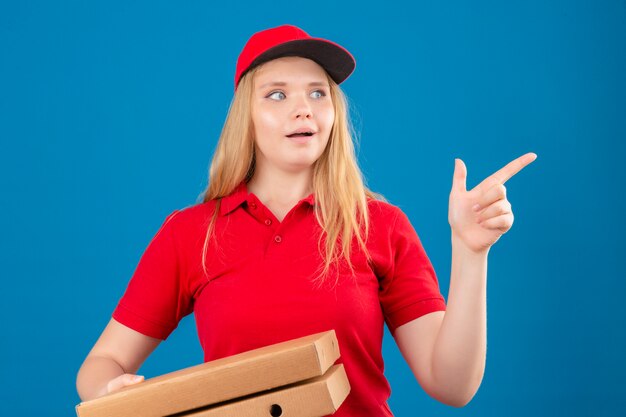 Giovane donna delle consegne che indossa la maglietta polo rossa e cappuccio in piedi con scatole per pizza cercando sorpreso puntare il dito a lato su sfondo blu isolato