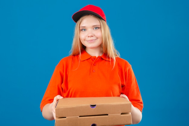 Giovane donna delle consegne che indossa maglietta polo arancione e cappuccio rosso che tiene scatole per pizza guardando la fotocamera con il sorriso sul viso isolato su sfondo blu