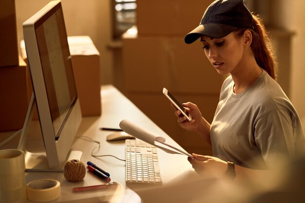 Giovane donna delle consegne che utilizza il telefono cellulare mentre esamina la lista di spedizione in ufficio