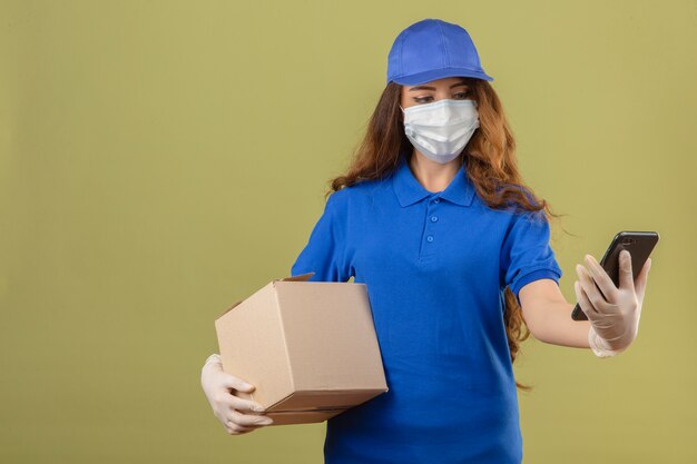 Giovane donna delle consegne con i capelli ricci che indossa la maglietta polo blu e berretto in maschera protettiva medica in piedi con la scatola di cartone e guardando lo schermo dello smartphone con la faccia seria sopra verde isolato