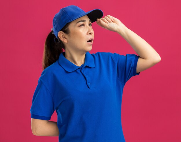 Giovane donna delle consegne in uniforme blu e berretto che guarda da parte con un'espressione pensierosa che pensa in piedi sul muro rosa pink