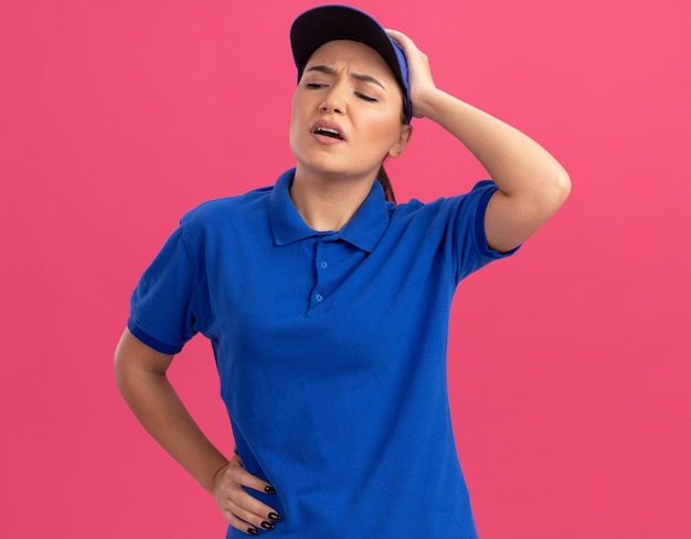 Giovane donna delle consegne in uniforme blu e berretto che sembra confuso con la mano sulla sua testa per errore in piedi sopra il muro rosa