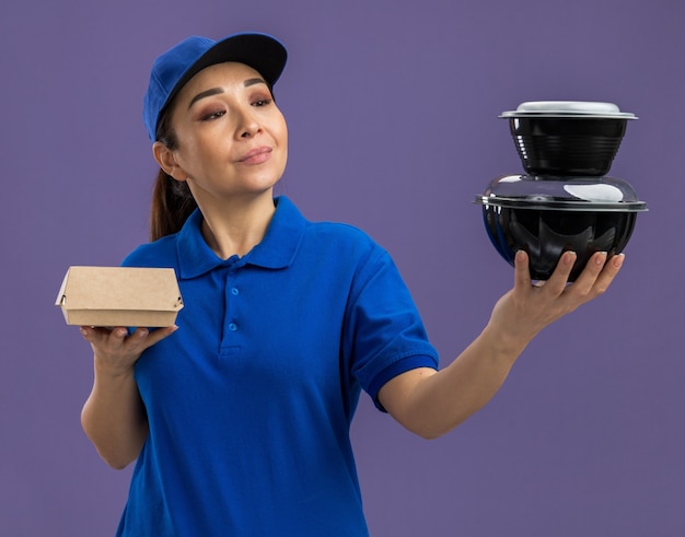 Giovane donna delle consegne in uniforme blu e berretto che tiene in mano pacchi di cibo guardandoli con un sorriso sul viso in piedi sul muro viola purple
