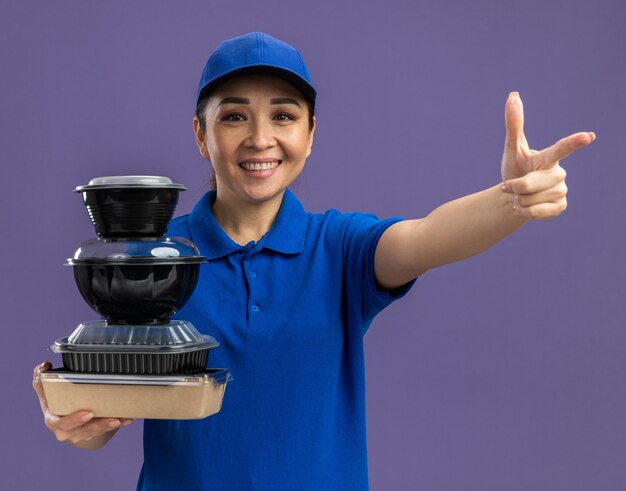 Giovane donna delle consegne in uniforme blu e berretto che tiene una pila di pacchi di cibo che sorride allegramente indicando con il dito indice in piedi sul muro viola