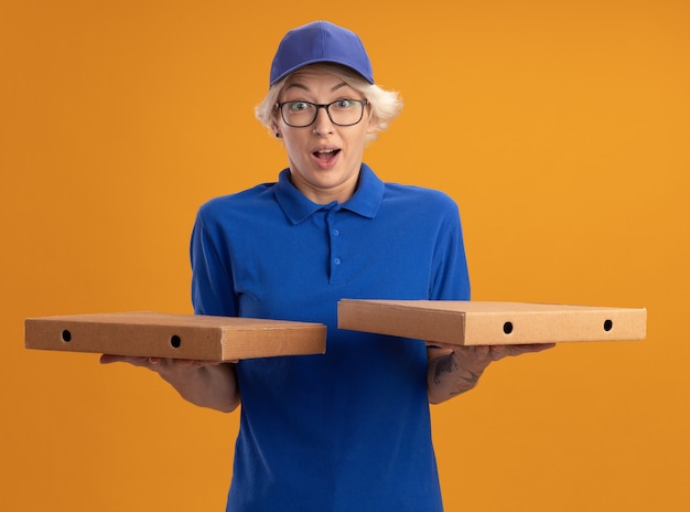 Giovane donna delle consegne in uniforme blu e berretto con gli occhiali in possesso di scatole per pizza stupita e sorpresa sulla parete arancione