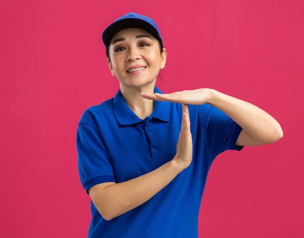 Giovane donna delle consegne in uniforme blu e berretto con un sorriso sul viso che fa un gesto di pausa con le mani