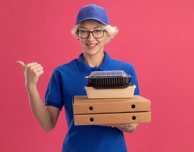 Giovane donna delle consegne in uniforme blu e cappello con gli occhiali che tengono le scatole della pizza e confezioni di cibo sorridendo allegramente indicando con il dito indice di lato sopra il muro rosa