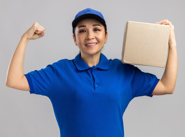 Giovane donna delle consegne in uniforme blu e cappuccio che tiene in mano una scatola di cartone felice ed eccitata che alza il pugno sorridendo allegramente in piedi sul muro bianco