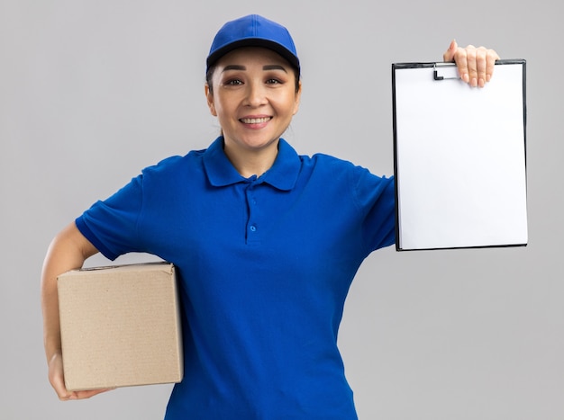 Giovane donna delle consegne in uniforme blu e cappuccio che tiene una scatola di cartone che mostra appunti con pagine bianche sorridenti fiduciosi in piedi sul muro bianco