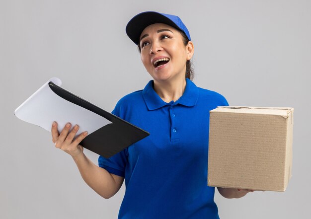 Giovane donna delle consegne in uniforme blu e cappuccio con scatola di cartone e appunti che guarda da parte sorridendo allegramente in piedi sul muro bianco