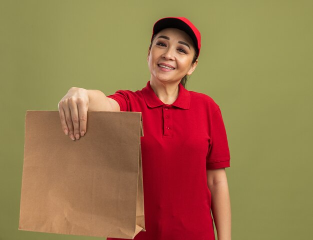 Giovane donna delle consegne in uniforme rossa e berretto che tiene in mano un pacchetto di carta con un sorriso sul viso felice e positivo in piedi sul muro verde green