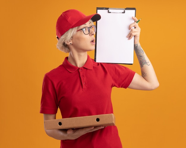 Giovane donna delle consegne in uniforme rossa e berretto con gli occhiali che tengono la scatola della pizza che mostra la lavagna per appunti guardandolo incuriosito in piedi sopra la parete arancione