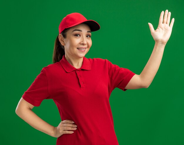 Giovane donna delle consegne in uniforme rossa e berretto felice e positivo che alza la mano sorridendo allegramente in piedi sul muro verde green