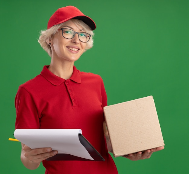 Giovane donna delle consegne in uniforme rossa e cappello con gli occhiali con la scatola di cartone che tiene appunti con pagine vuote e matita che sorride allegramente in piedi sopra la parete verde