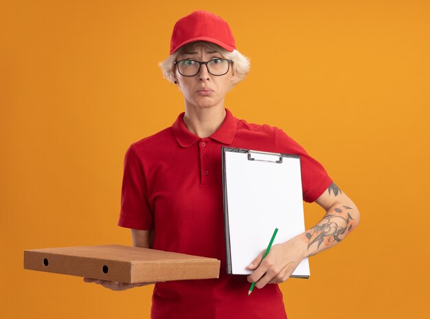 Giovane donna delle consegne in uniforme rossa e cappello con gli occhiali tenendo la scatola della pizza e appunti con la matita che sembra confuso e molto ansioso in piedi sopra la parete arancione