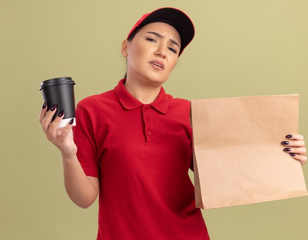 Giovane donna delle consegne in uniforme rossa e cappuccio che tiene il pacchetto di carta e la tazza di caffè guardando la parte anteriore dispiaciuta in piedi sopra la parete verde