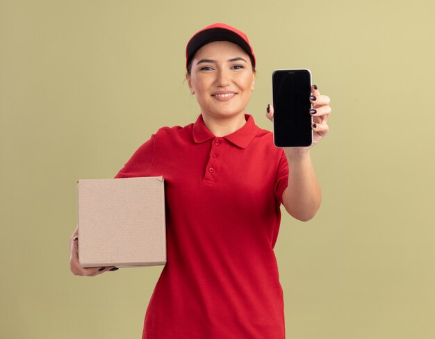 Giovane donna delle consegne in uniforme rossa e cappuccio che tiene la scatola di cartone che mostra smartphone guardando davanti con il sorriso sul viso in piedi sopra la parete verde