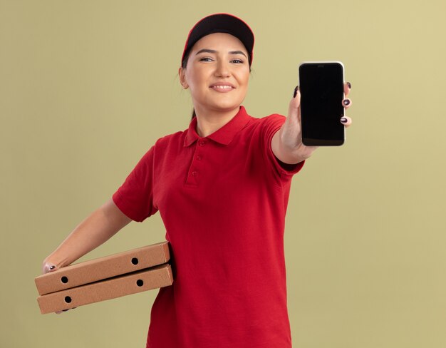 Giovane donna delle consegne in uniforme rossa e cappuccio che tiene le scatole per pizza che mostrano il telefono cellulare guardando la parte anteriore sorridente allegramente in piedi sopra la parete verde