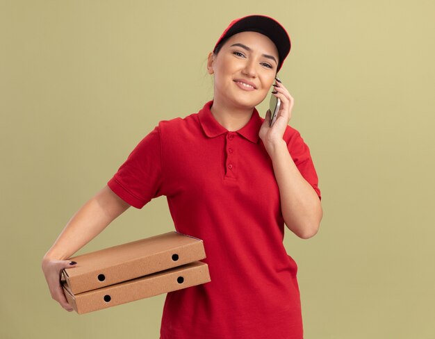 Giovane donna delle consegne in uniforme rossa e cappuccio che tiene le scatole per pizza sorridente con la faccia felice mentre parla al telefono cellulare in piedi sopra la parete verde