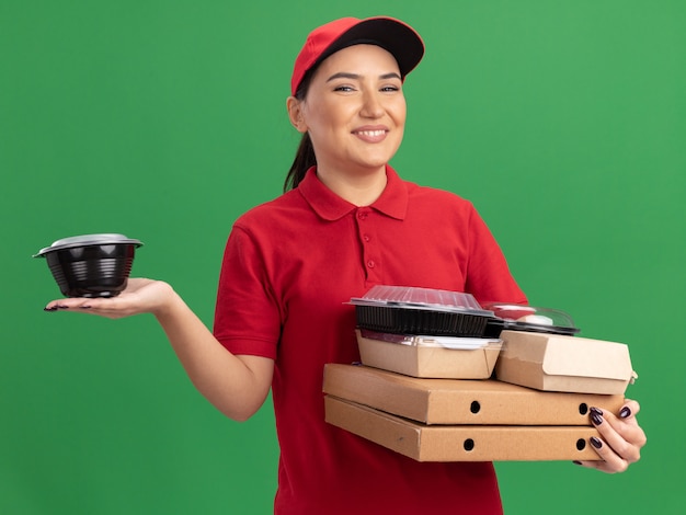 Giovane donna delle consegne in uniforme rossa e cappuccio che tiene scatole di pizza e pacchetti di cibo guardando la parte anteriore sorridente allegramente in piedi sopra la parete verde