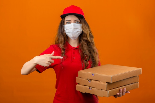 Giovane donna delle consegne indossando maglietta polo rossa e cappuccio in maschera protettiva medica in piedi con una pila di scatole e puntare il dito indice verso di loro guardando la fotocamera con faccia seria sopra oran isolato