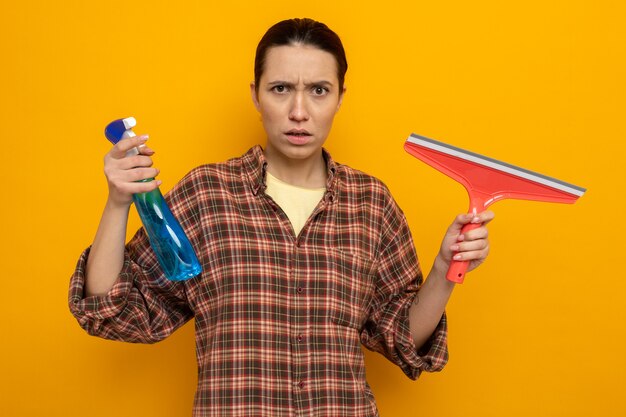 Giovane donna delle pulizie in abiti casual che tiene in mano uno spray detergente e una scopa che guarda con faccia seria