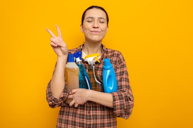 Giovane donna delle pulizie in abiti casual in possesso di forniture per la pulizia felice e allegra che mostra il segno a V in piedi sul muro arancione