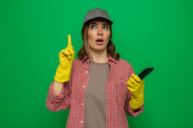 Giovane donna delle pulizie in camicia a quadri e berretto che indossa guanti di gomma che tengono lo smartphone alzando lo sguardo mostrando il dito indice che ha una nuova idea in piedi su sfondo verde