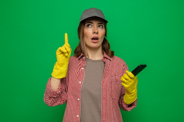 Giovane donna delle pulizie in camicia a quadri e berretto che indossa guanti di gomma che tengono lo smartphone alzando lo sguardo mostrando il dito indice che ha una nuova idea in piedi su sfondo verde