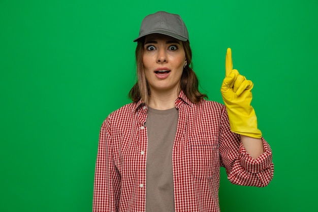 Giovane donna delle pulizie in camicia a quadri e berretto che indossa guanti di gomma guardando la telecamera sorpresa mostrando il dito indice con una nuova idea in piedi su sfondo verde