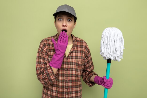 Giovane donna delle pulizie in camicia a quadri e berretto in guanti di gomma che tiene mop guardando davanti stupito e scioccato che copre la bocca con la mano in piedi sul muro verde