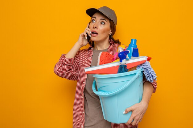 Giovane donna delle pulizie in camicia a quadri e cappello che tiene il secchio con strumenti per la pulizia che sembra sorpresa mentre parla al telefono cellulare in piedi su sfondo arancione orange