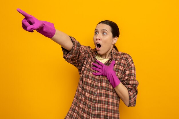 Giovane donna delle pulizie in camicia a quadri in guanti di gomma che guarda da parte stupita e sorpresa indicando con il dito indice qualcosa in piedi sul muro arancione