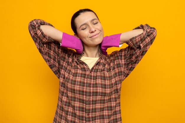 Giovane donna delle pulizie in camicia a quadri in guanti di gomma felice e contenta che si allunga in piedi sul muro arancione