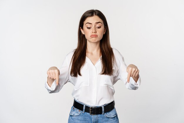 Giovane donna delusa che punta il dito verso il basso, guardando sotto con rammarico e tristezza, in piedi in camicetta e jeans su sfondo bianco