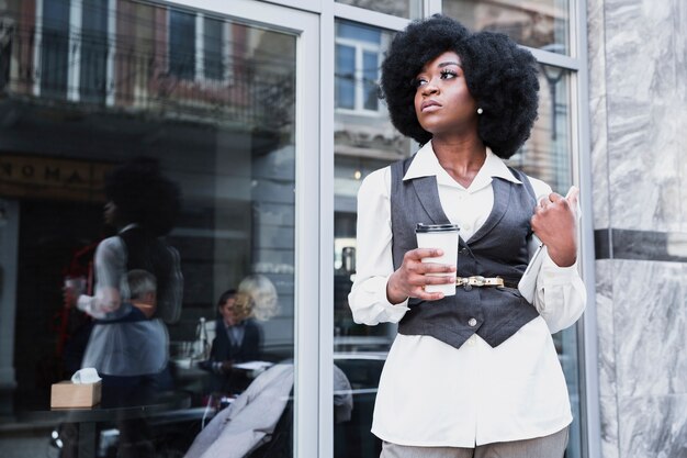 Giovane donna di affari africana alla moda che tiene la tazza di caffè asportabile e distogliere lo sguardo digitale della compressa