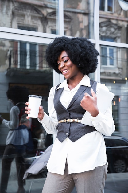 Giovane donna di affari africana sorridente che tiene la tazza di caffè asportabile e la compressa digitale a disposizione