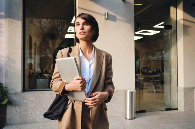 Giovane donna di affari alla moda attraente con il computer portatile che distoglie lo sguardo sognante sulla via della città