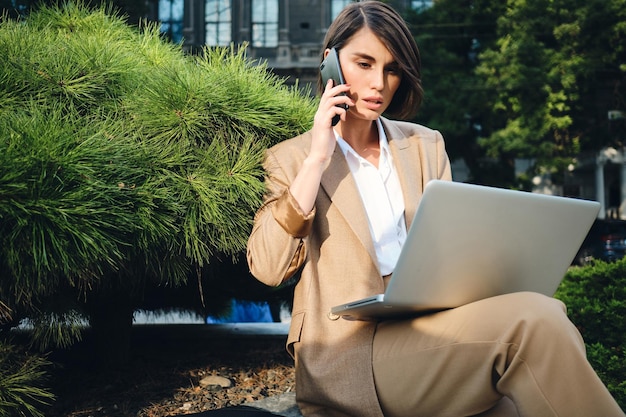 Giovane donna di affari alla moda seria che parla sul cellulare e che lavora al computer portatile all'aperto