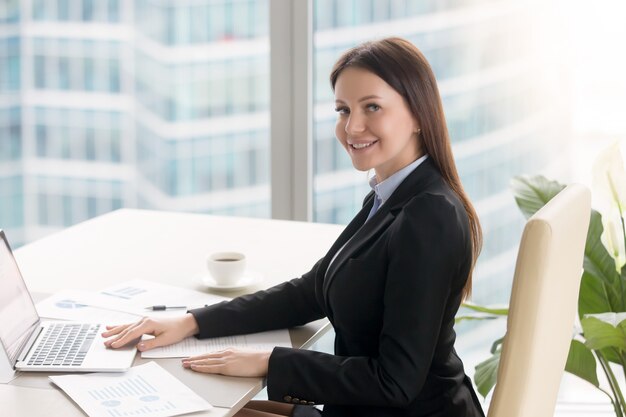 Giovane donna di affari allegra sorridente che lavora alla scrivania con il computer portatile