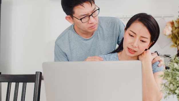 Giovane donna di affari asiatica seria, stress, stanca e malata mentre si lavora al computer portatile a casa. Il marito la consola mentre lavora sodo nella cucina moderna a casa la mattina.