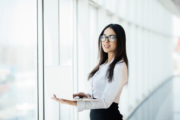 Giovane donna di affari che lavora nel suo ufficio di lusso in possesso di un computer portatile in piedi contro la finestra panoramica con vista sul quartiere degli affari