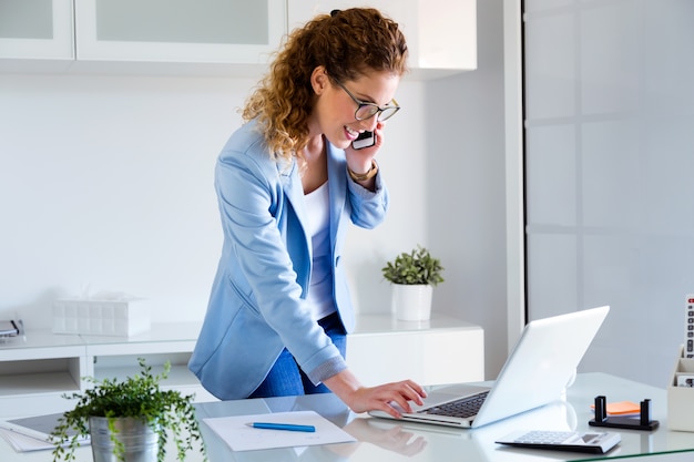 Giovane donna di affari che parla sul telefono cellulare mentre usando il suo computer portatile nell&#39;ufficio.