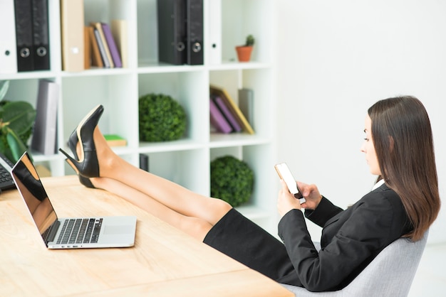 Giovane donna di affari che si siede sulla sedia con le gambe incrociate sullo scrittorio facendo uso dello smartphone