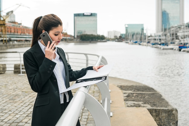 Giovane donna di affari che sta al porto che parla sul telefono cellulare mentre esaminando lavagna per appunti