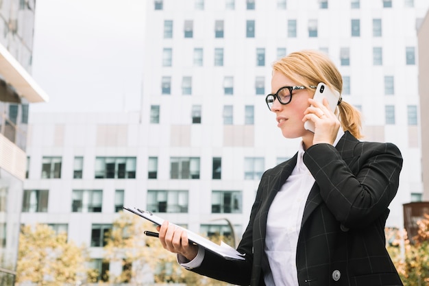 Giovane donna di affari che sta fuori degli edifici corporativi che parla sul telefono cellulare