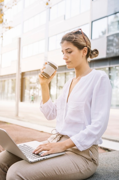 Giovane donna di affari che tiene la tazza di caffè asportabile che scrive sul computer portatile