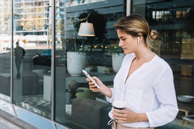 Giovane donna di affari che tiene la tazza di caffè eliminabile facendo uso del telefono cellulare