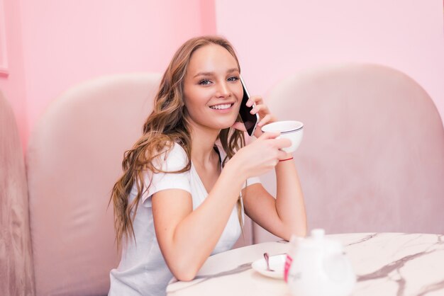 Giovane donna di affari in vestito grigio che si siede al tavolo in caffè, parlando sul telefono cellulare mentre si prendono appunti in notebook