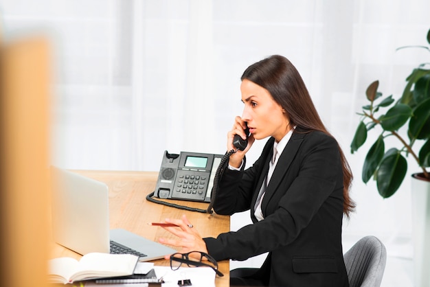 Giovane donna di affari seria che si siede allo scrittorio che comunica sul telefono